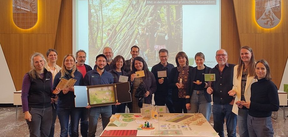 Treffen der AG Bildung für nachhaltige Entwicklung Naturpark Saar-Hunsrück Teilnehmer des Treffens der AG Bildung für nachhaltige Entwicklung Naturpark Saar-Hunsrück stehen vor einem Foto das Kinder im Wald zeigt. Im Vordergrund ein Tisch mit den neu gestalteten Spielen für Kindergärten und Schulen.