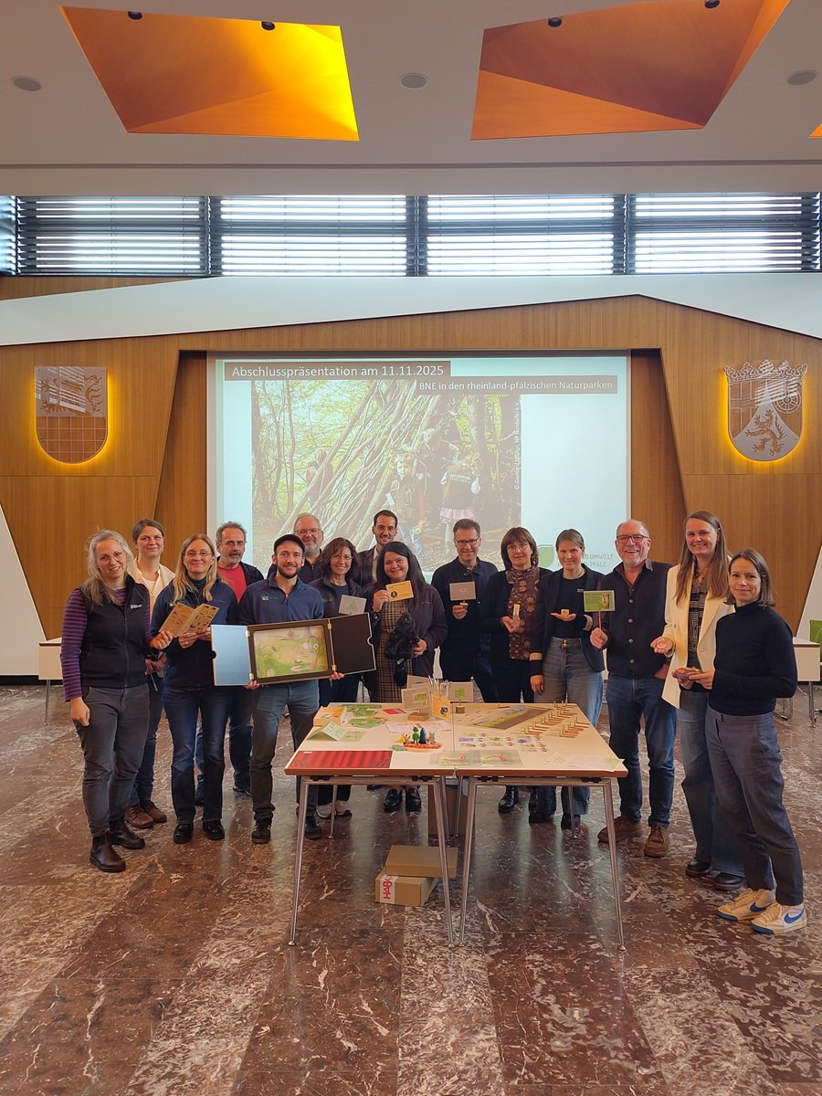 Treffen der AG Bildung für nachhaltige Entwicklung Naturpark Saar-Hunsrück Teilnehmer des Treffens der AG Bildung für nachhaltige Entwicklung Naturpark Saar-Hunsrück stehen vor einem Foto das Kinder im Wald zeigt. Im Vordergrund ein Tisch mit den neu gestalteten Spielen für Kindergärten und Schulen.