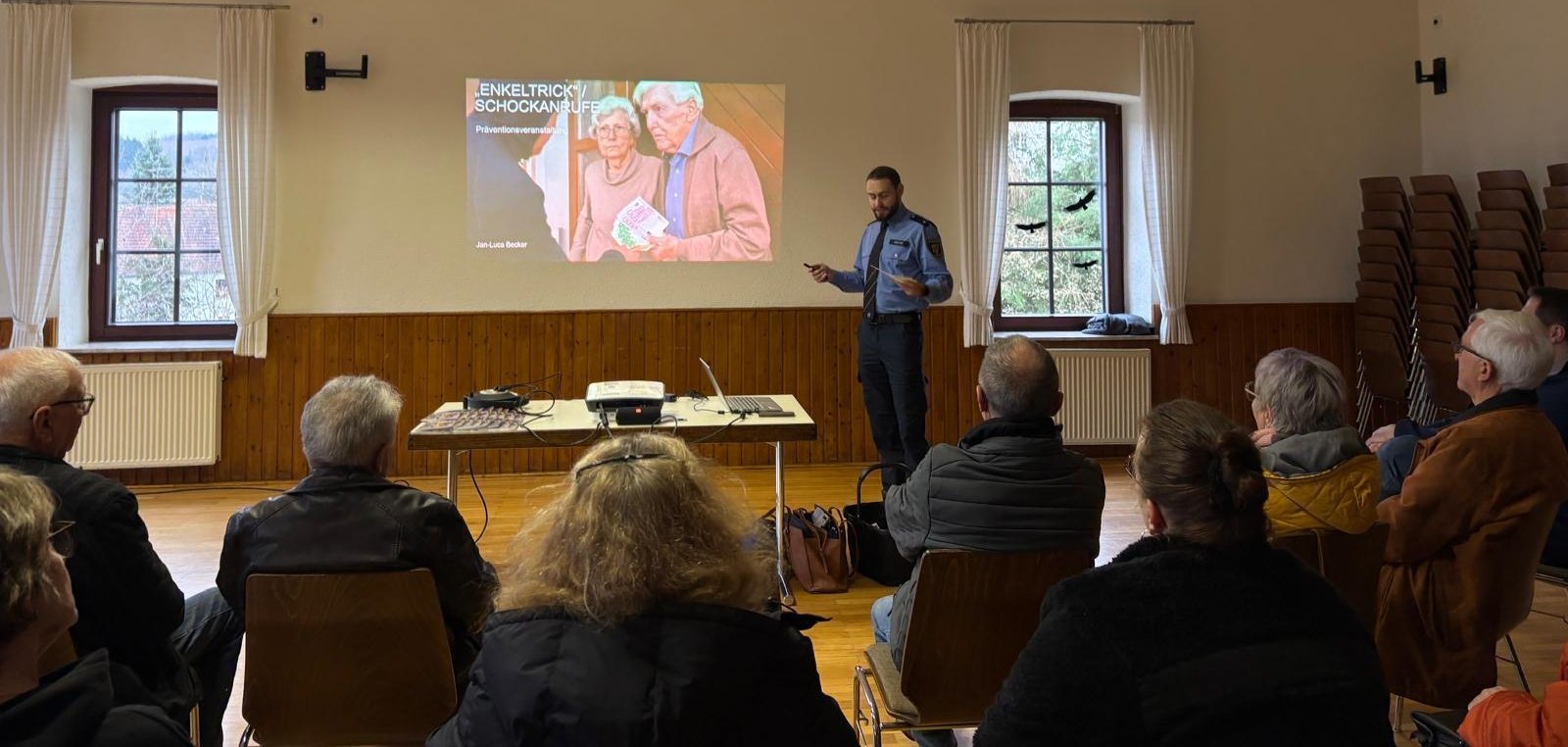 Vortrag der Polizei zur Prävention bei Schockanrufen. Ältere Personen sitzen in einem Saal. Vorne steht ein Polizeibeamter vor einer Leinwand.