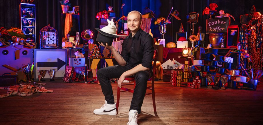Zauberkünstler Jakob Mathias Zauberkünstler Jakob Mathias sitzt auf der Bühne. in der Hand hält er einen Zylinder aus dem ein weißes Kaninchen herausschaut.