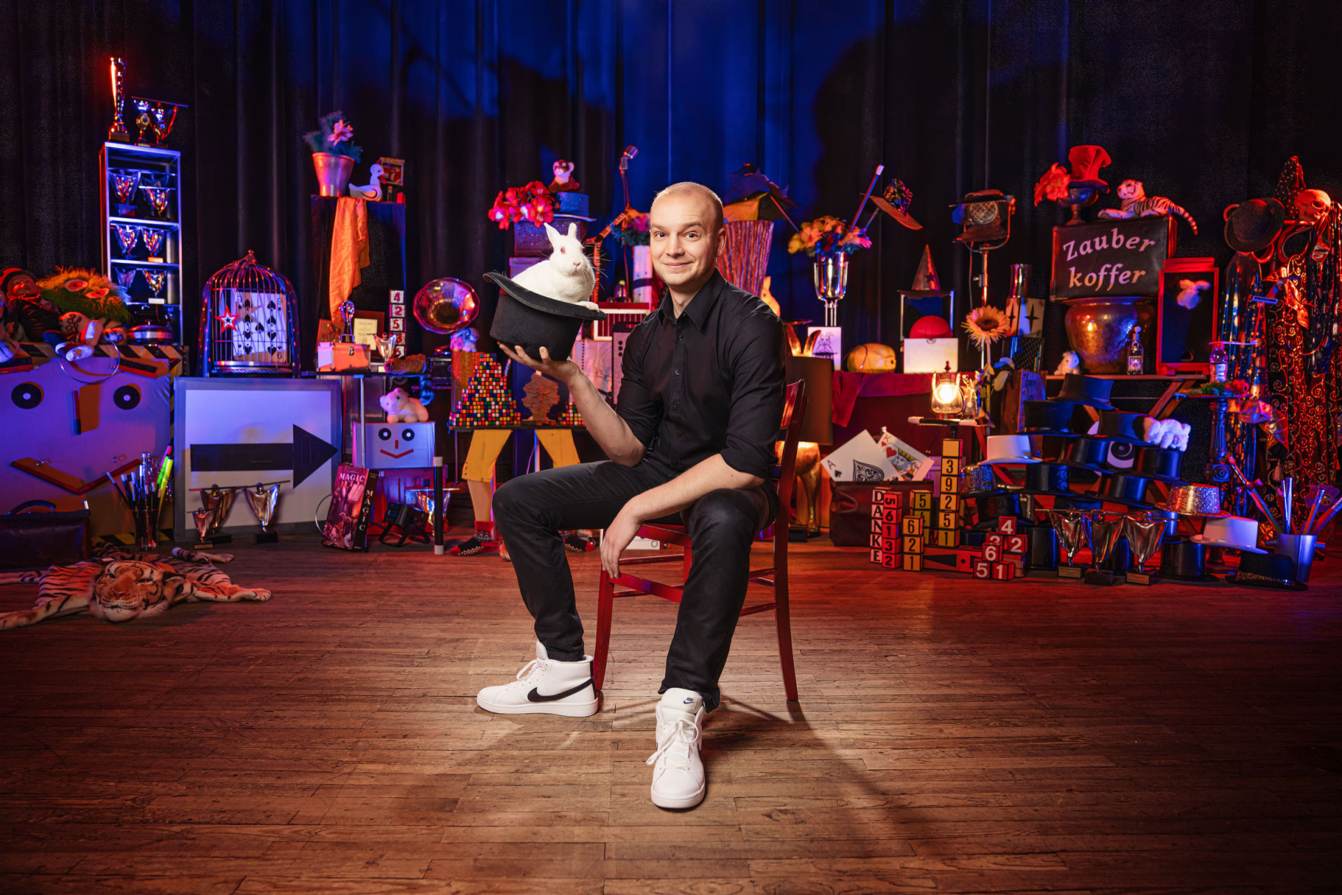 Zauberkünstler Jakob Mathias sitzt auf der Bühne. in der Hand hält er einen Zylinder aus dem ein weißes Kaninchen herausschaut.