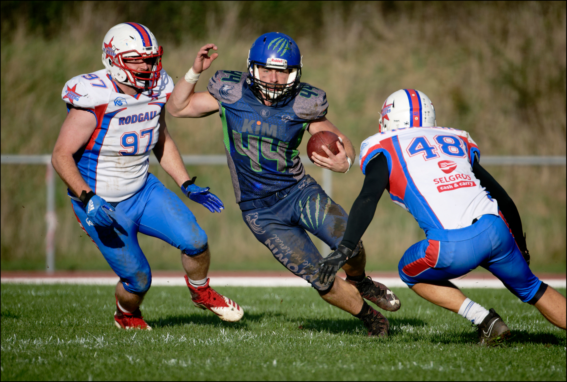 Drei American Footbal Spieler in einer Spielsituation. Die mittlere Person hat den Ball. Gegnerische Spieler kommen von rechts und links auf ihn zu.