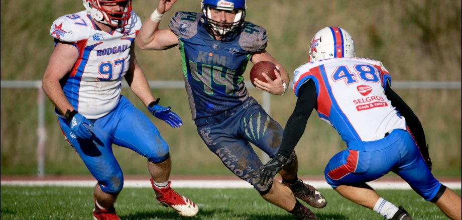 American Footbal Spieler Drei American Footbal Spieler in einer Spielsituation. Die mittlere Person hat den Ball. Gegnerische Spieler kommen von rechts und links auf ihn zu.