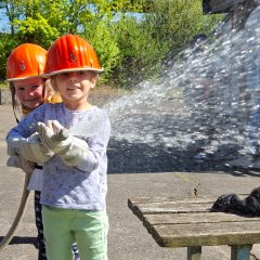 Zwei Jungs spritzen mit einem Feuerwehrschlauch. Sie tragen Helme und freuen sich.