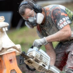 Ein Motorsägenkünstler am Bauernmarkt in Berglangenbach formt eine Schnecke aus Holz mit seiner Motorsäge. Er trägt eine Schutzbrille und eine Atemmaske. Die Holzspäne wirbeln durch die Luft.