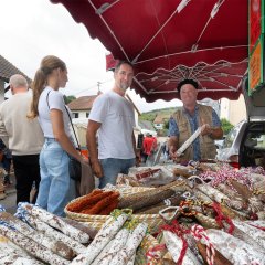 Salamistand auf dem Bauernmarkt in Berglangenbach. Auf dem Tisch liegen unterschiedliche Salamiwürste. Der Verkäufer hält eine Wurst in die Kamera. vor dem Stand lächelt ein Kunde in die Kamera.