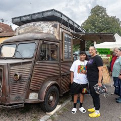 Waffeln- und Crepe-Stand auf dem Bauernmarkt in Berglangenbach. Der Stand ist ein Verkaufsauto, das wie ein alter Lieferwagen gestaltet ist. An der Seite ist eine große Klappe hochgeklappt, so dass man in den Wagen sehen kann. Davor stehen eine Frau und ein Junge und lächeln in die Kamera.